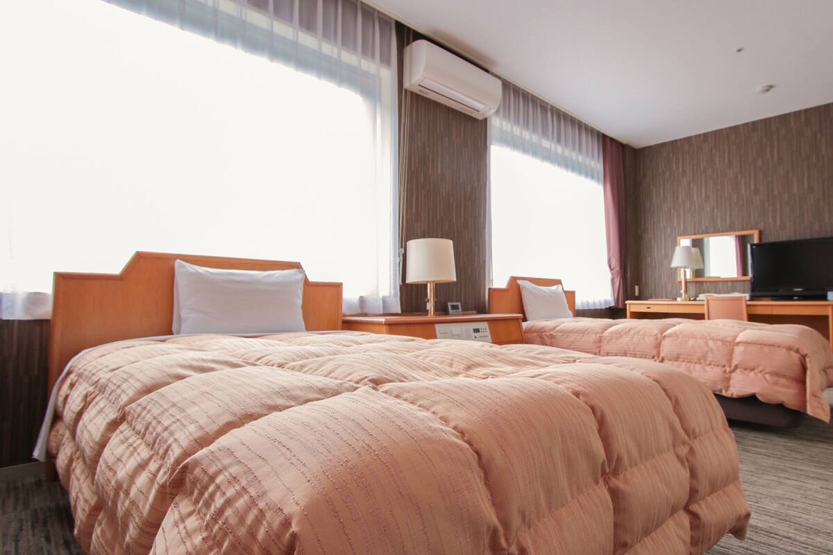 A hotel room with two beds, a TV, a desk, and a window with curtains.