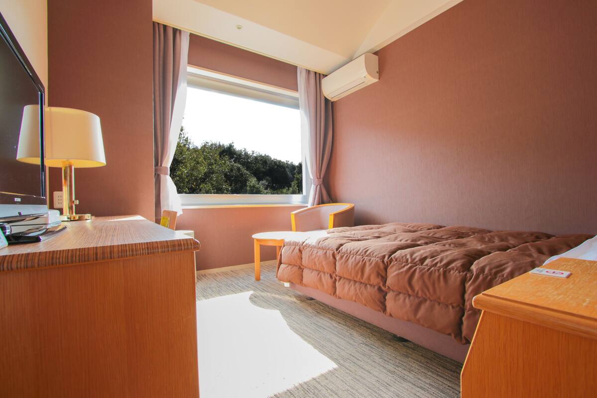 A hotel room with a bed, a desk, a chair, a window with curtains, and an air conditioning unit.