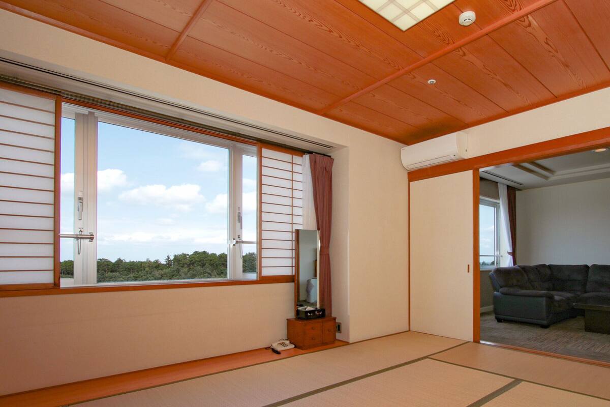 A room with tatami flooring, a sliding door leading to a living area with a sofa and a coffee table, and a large window with a view of trees.