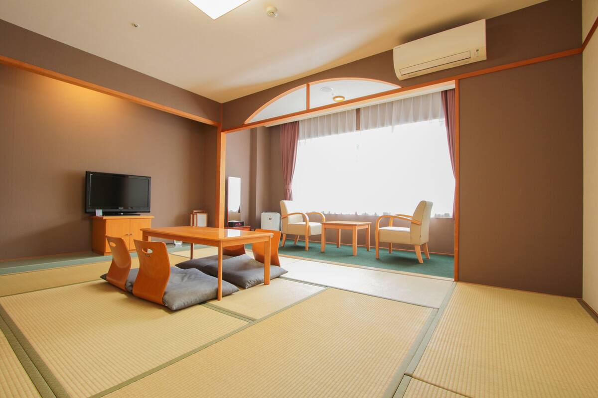 A room with tatami flooring, a television, a dining table with chairs, and a large window with curtains.