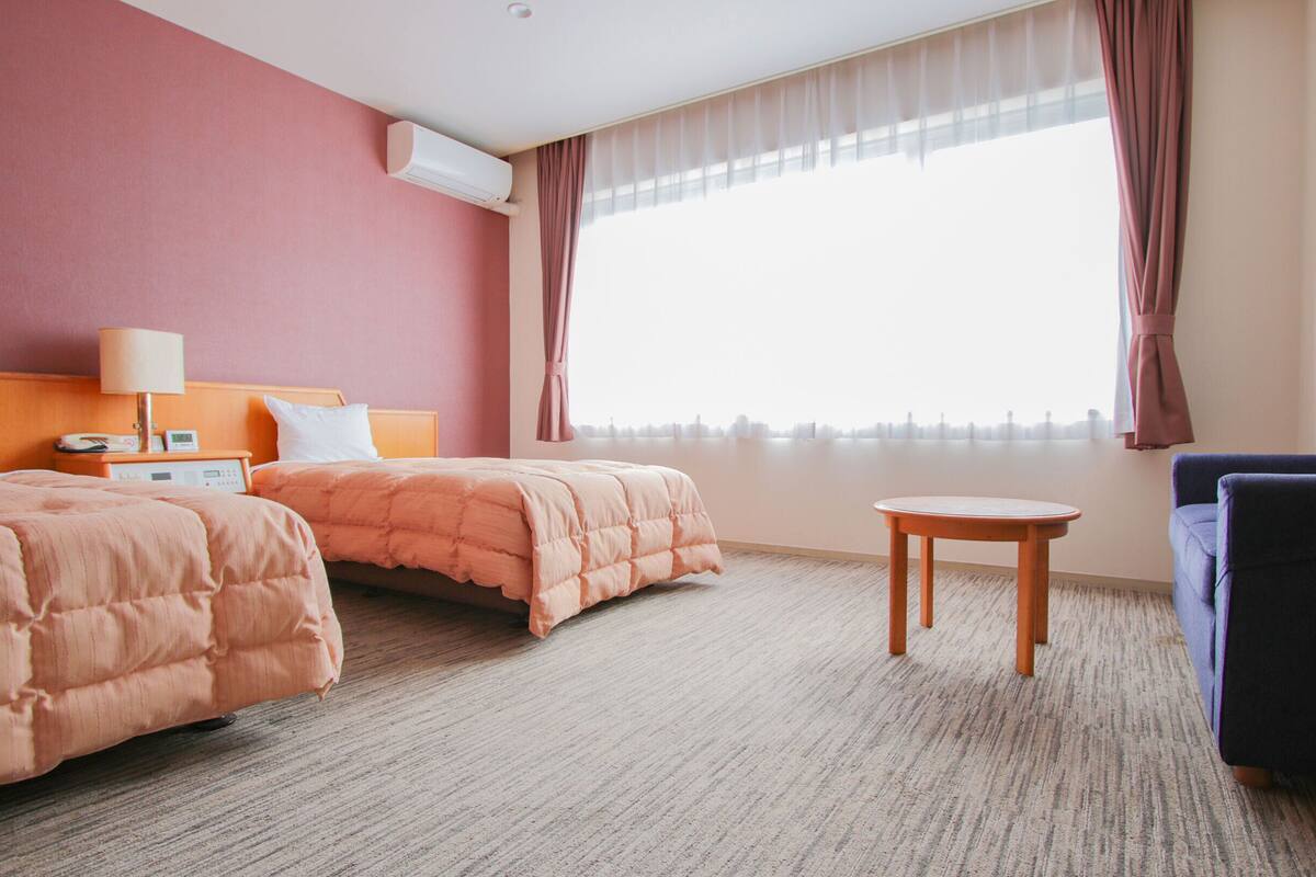 A hotel room with two beds, a small table, a sofa, a lamp, and an air conditioning unit.