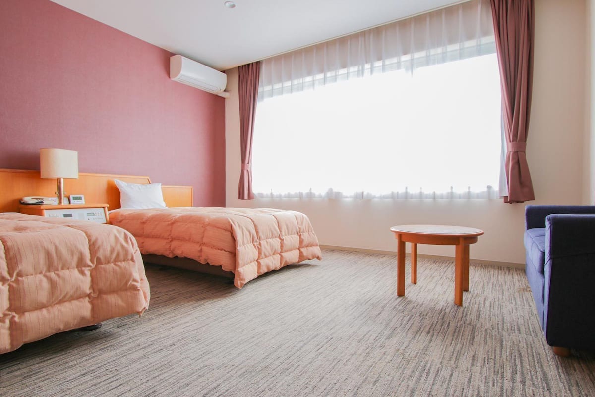 A hotel room with two beds, a small table, a sofa, a lamp, and an air conditioning unit.