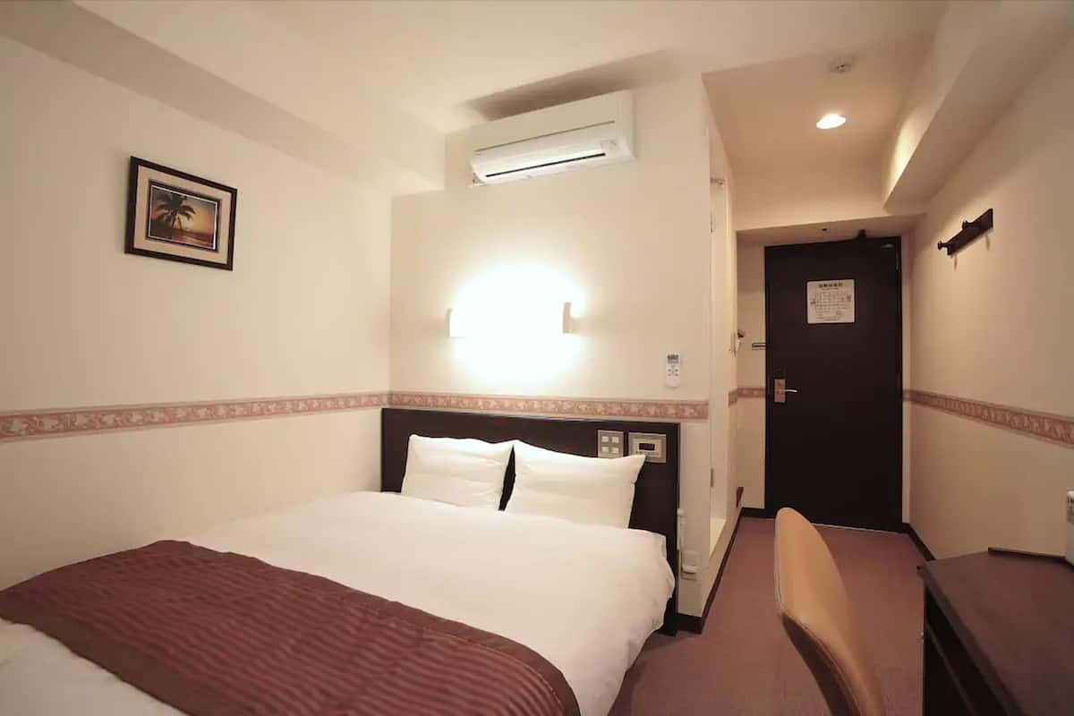 A hotel room with a bed, a bedside table, an air conditioning unit, a wall-mounted light, and a door with a sign.