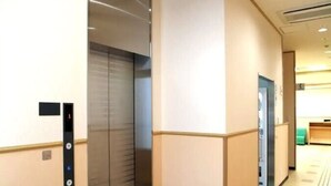 Un couloir avec un ascenseur, des murs de couleur beige et une porte donnant sur une pièce où se trouvent un lit et une table de chevet.