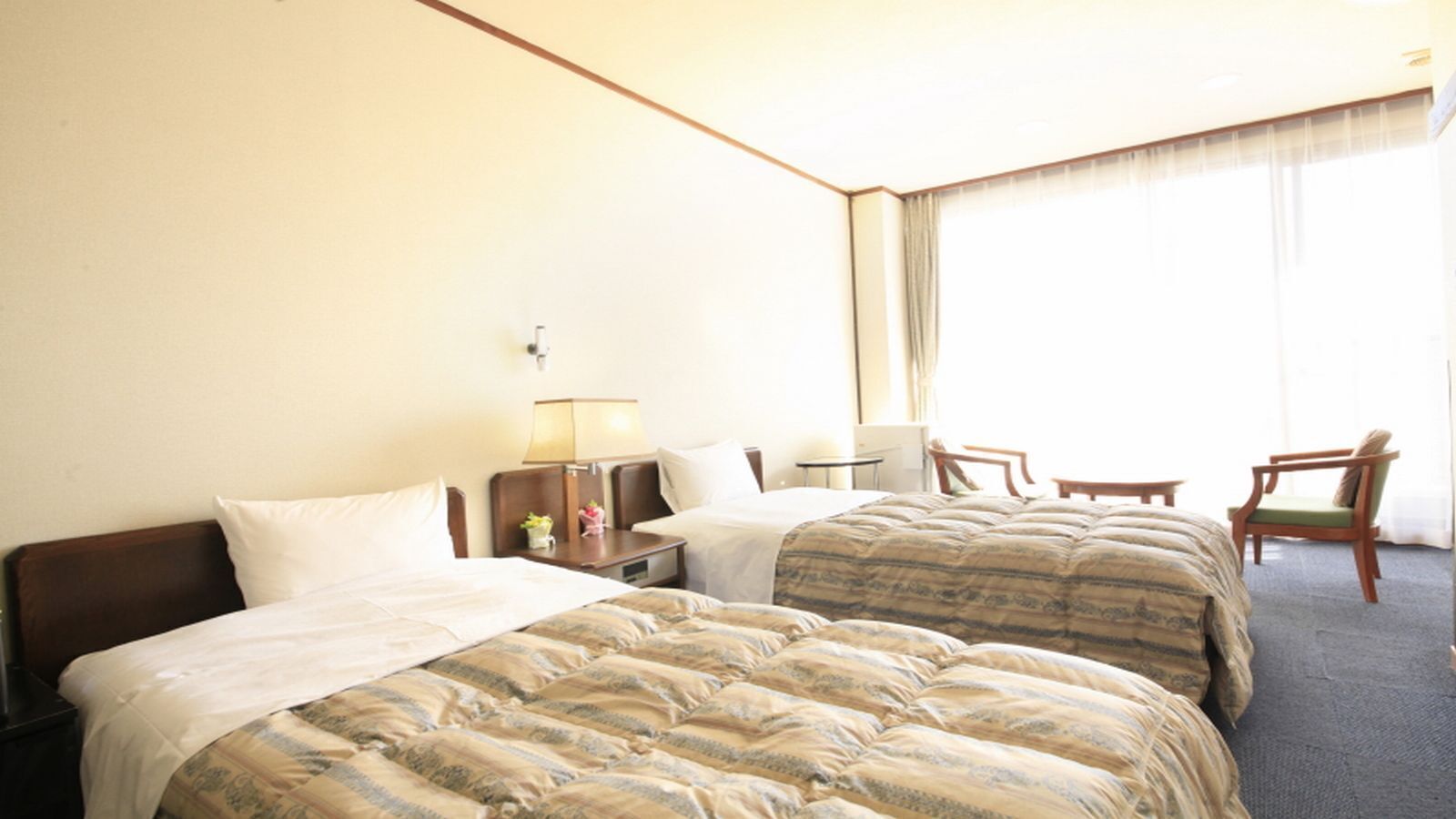 Una habitación de hotel con dos camas, una mesita de noche con lámpara, una silla y vistas al exterior a través de una ventana.