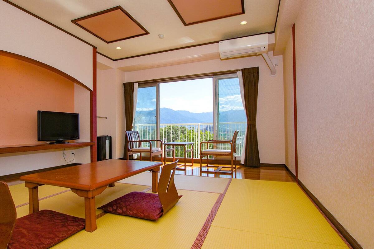 A hotel room with a TV, wooden furniture, tatami mats, a balcony with a mountain view.