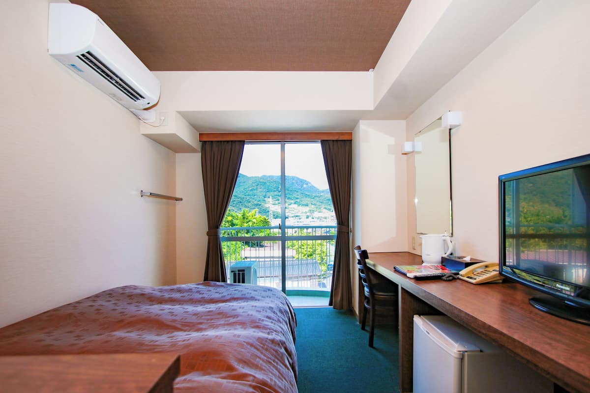 A hotel room with a bed, desk, and a television. There is a window with curtains, and an air conditioning unit is mounted on the wall.