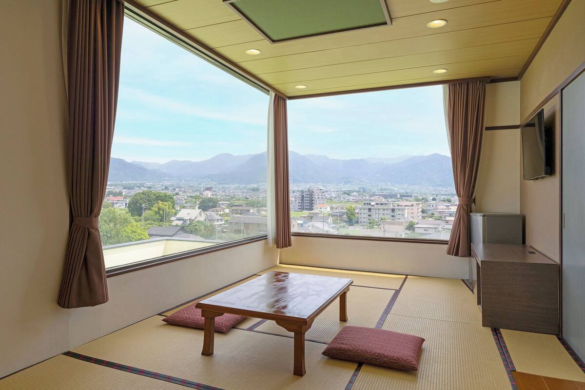 A room with tatami flooring, a wooden table, two cushions, a TV, and a view of a cityscape with mountains.