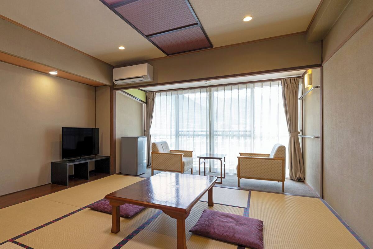 A room with tatami mats, a television, a refrigerator, and a view of the outside through large windows.