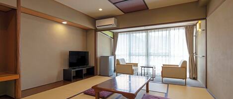 A room with tatami mats, a television, a refrigerator, and a view of the outside through large windows.