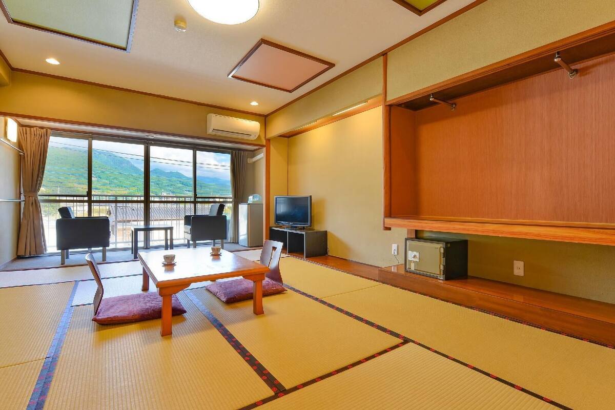 A traditional Japanese room with tatami flooring, a low table, chairs, a television, and a large window with a view of green hills.