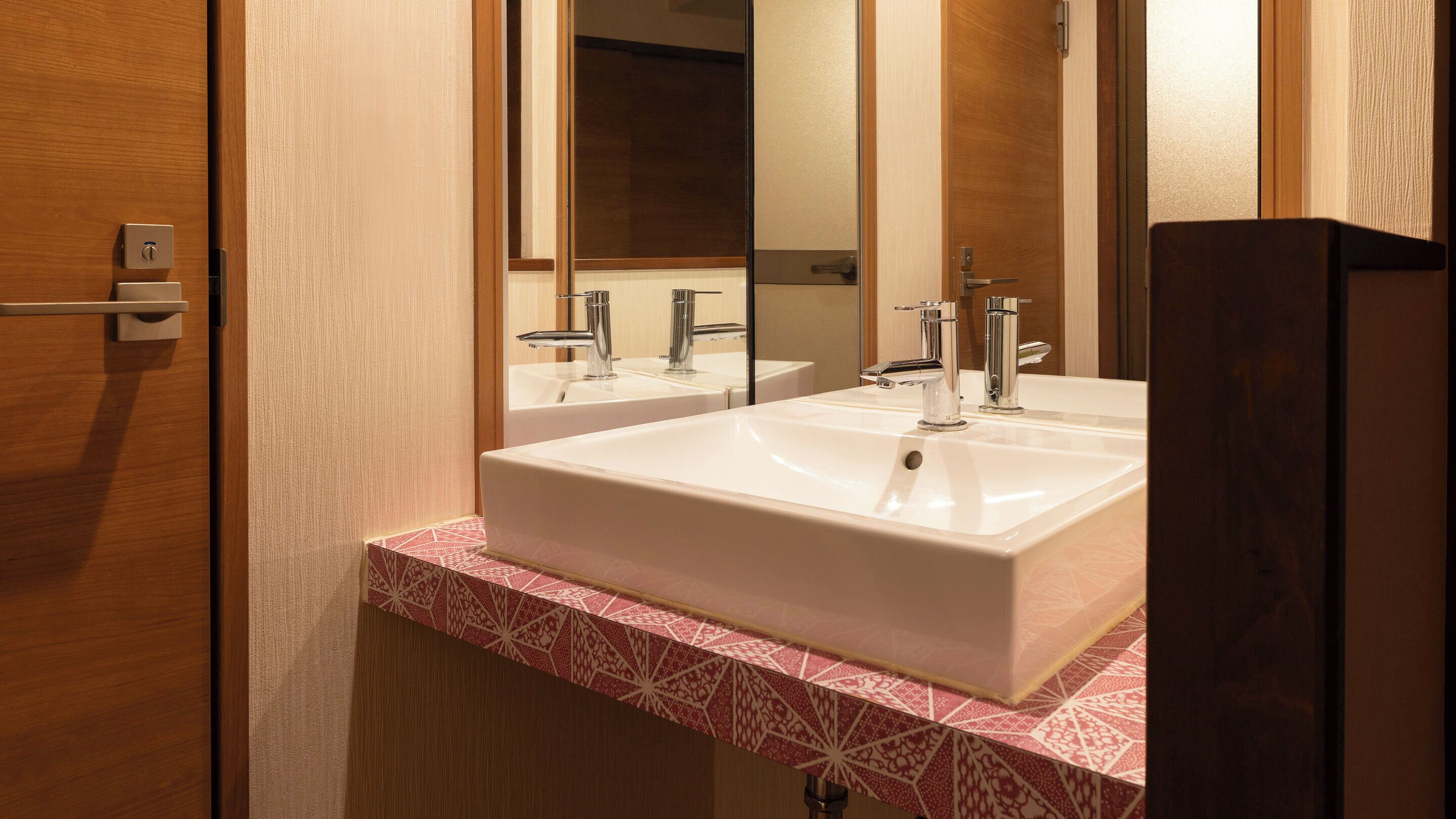 Un baño con lavamanos blanco, dos grifos, un espejo y una puerta con pomo.