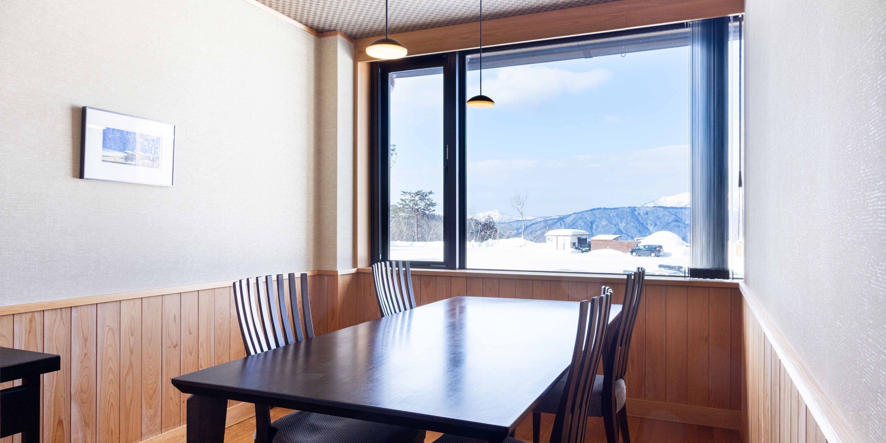 Un comedor con una mesa y sillas de madera, una ventana que ofrece vistas a colinas nevadas y un cuadro en la pared.