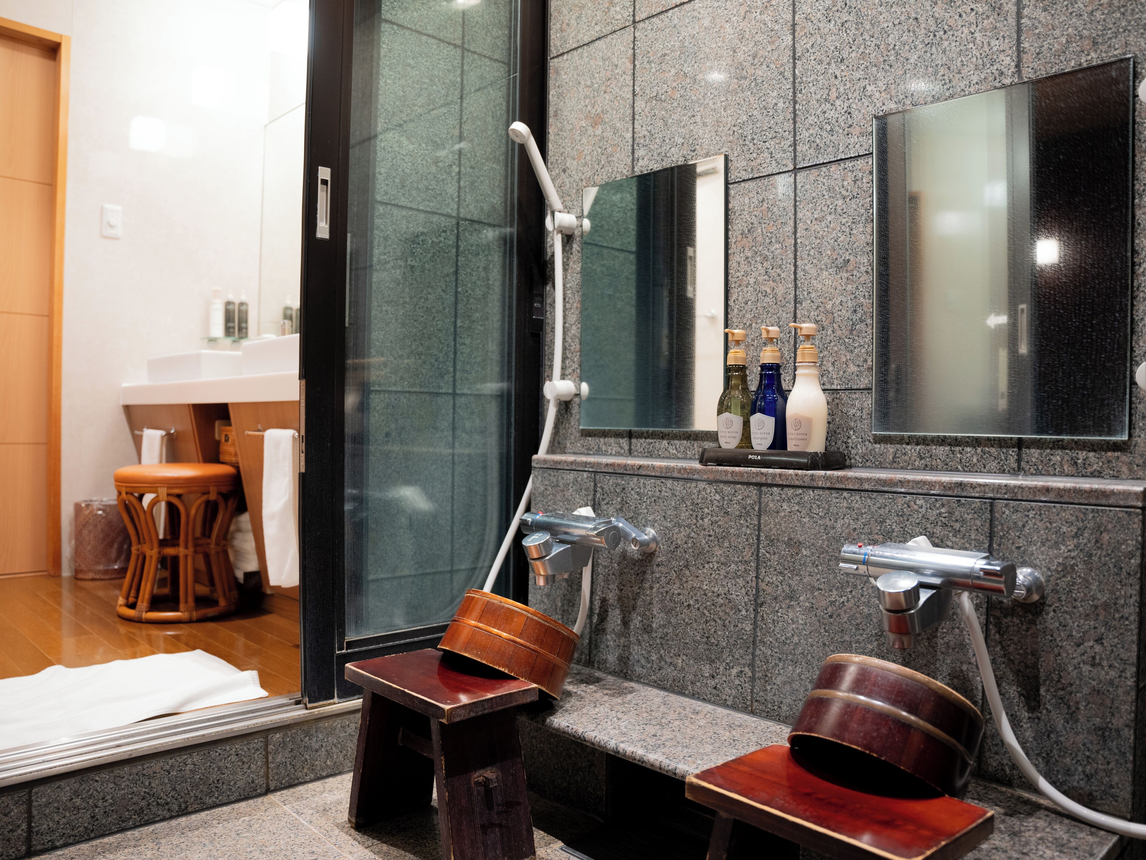 Un baño con cabina de ducha de vidrio, un taburete de madera, una mesita de madera y un espejo sobre el lavabo.