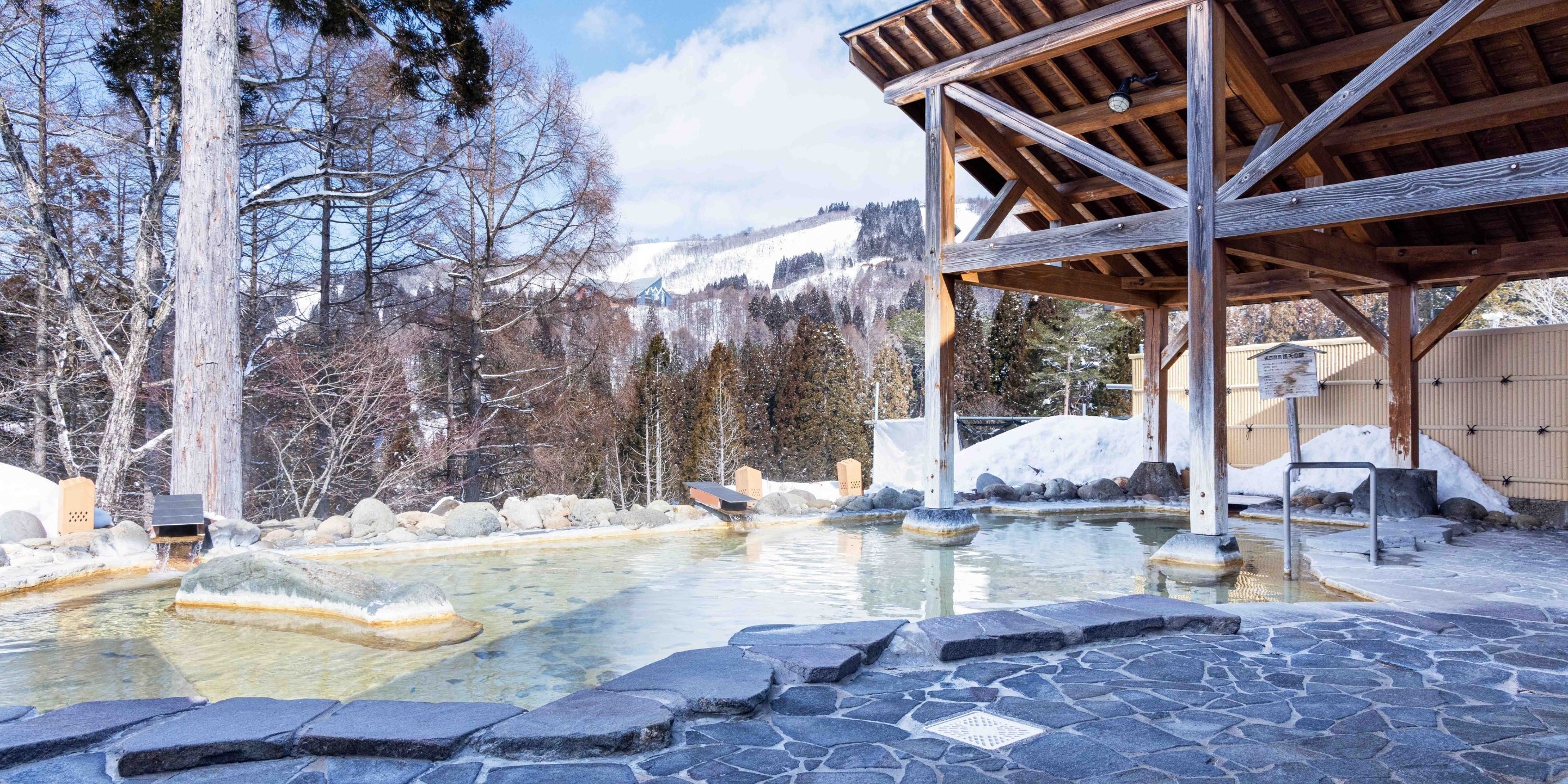 A wooden pavilion with a stone floor, a hot spring pool, and a mountainous backdrop.