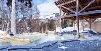 Un pabellón de madera con suelo de piedra, una piscina de aguas termales y montañas al fondo.