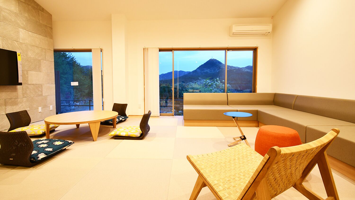 A modern living room with a sofa, a round table, and chairs, large windows with a mountain view, and a stone wall.