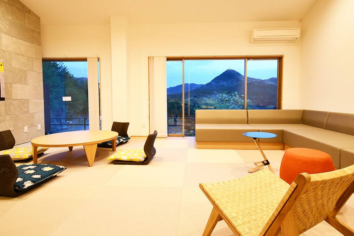 Un salon moderne avec un canapé, une table ronde et des chaises, de grandes fenêtres offrant une vue sur les montagnes et un mur en pierre.