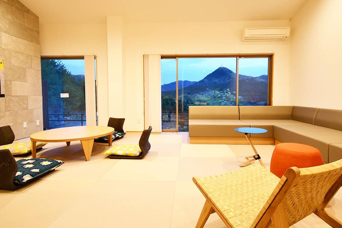 A modern living room with a sofa, a round table, and chairs, large windows with a mountain view, and a stone wall.