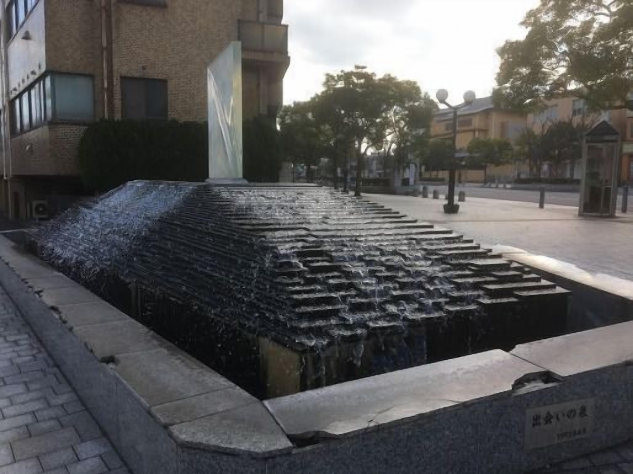 A water feature with a tiered design, surrounded by a paved area and buildings.