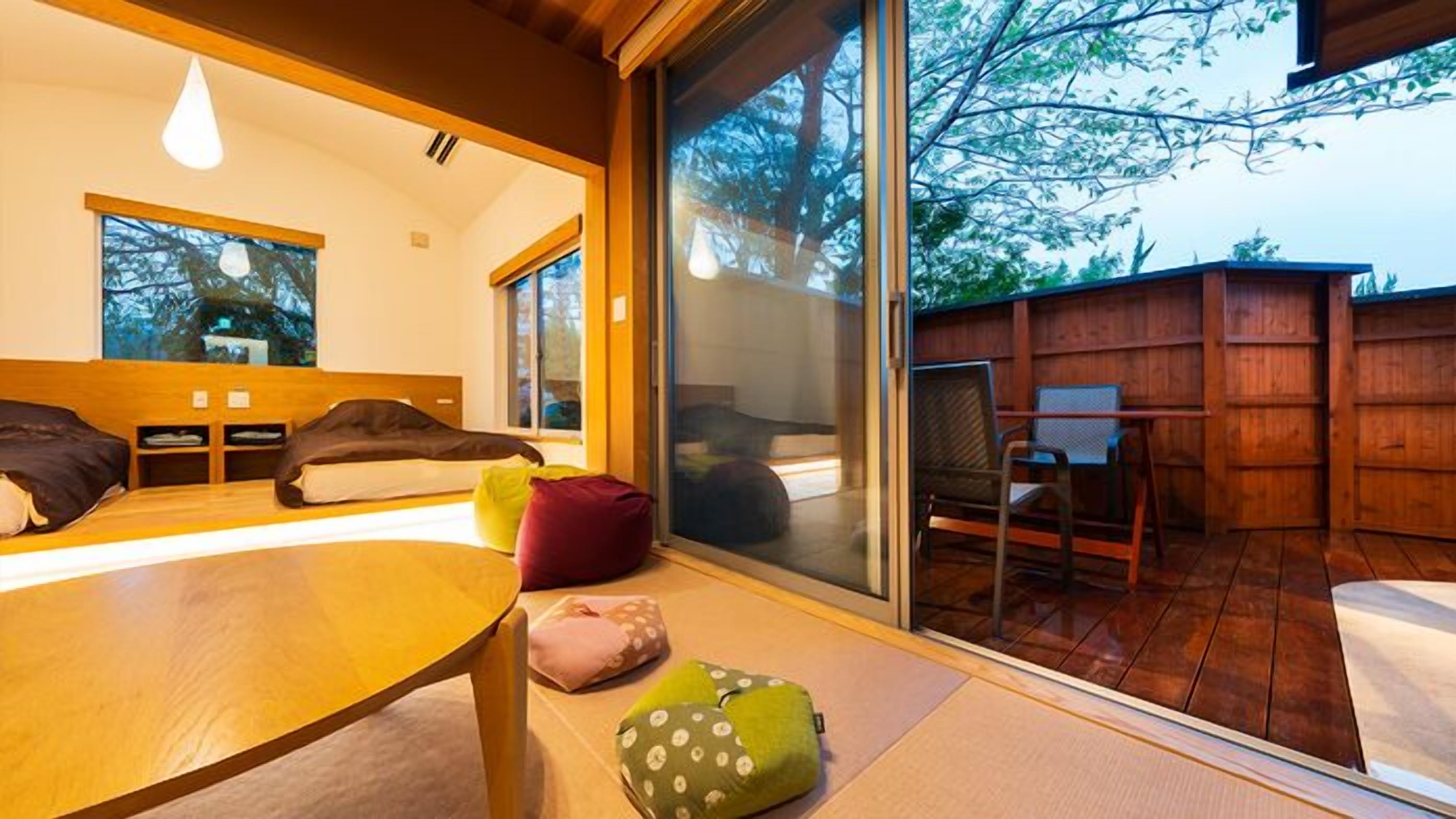 A modern room with a bed, a table, and a chair. There is a sliding glass door leading to an outdoor area with a wooden deck and a chair.