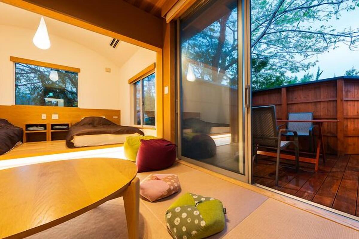 Une chambre moderne avec un lit, une table et une chaise. Une porte-fenêtre coulissante donne accès à un espace extérieur doté d’une terrasse en bois et d’une chaise.