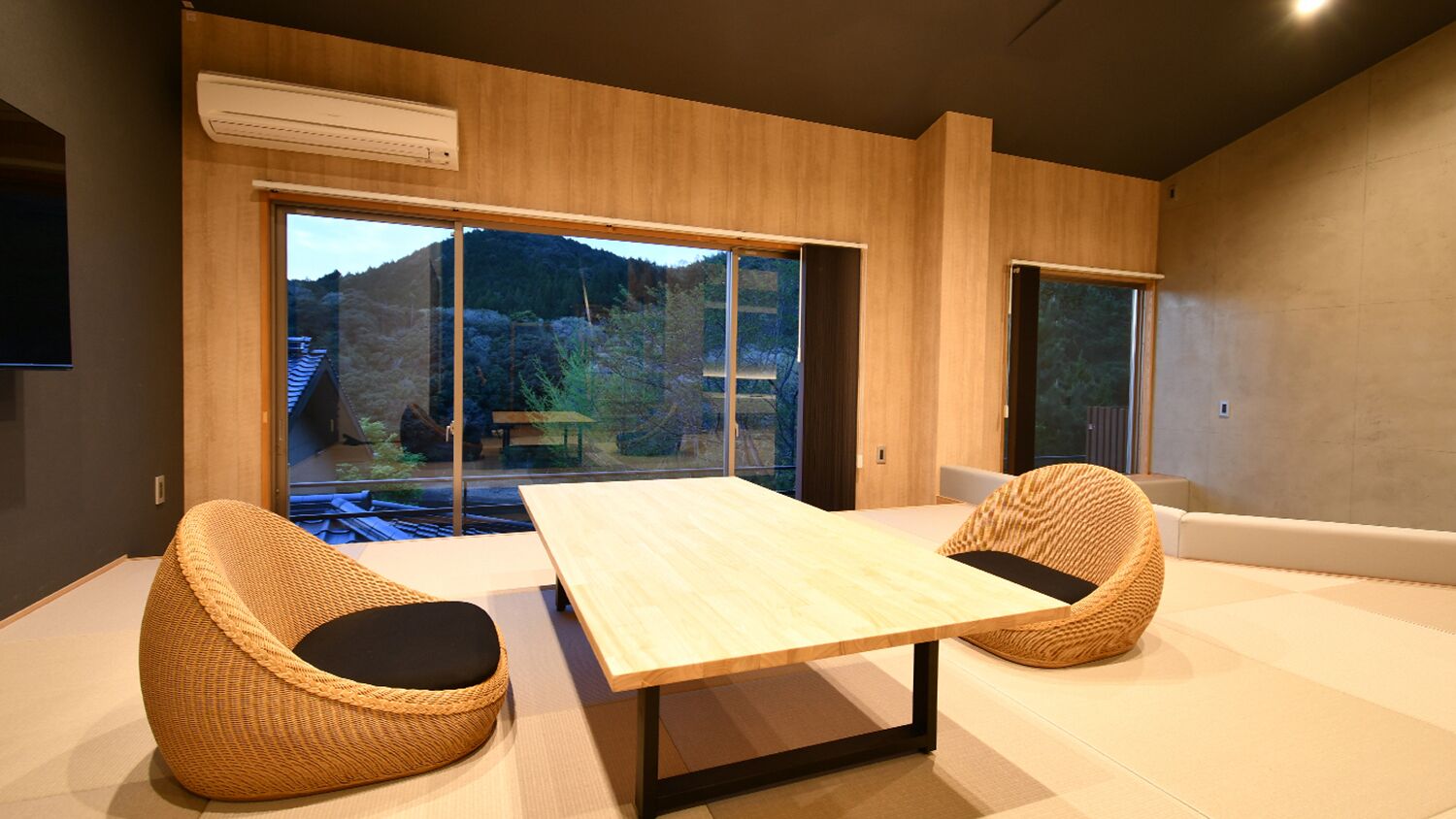 A modern room with a large table, two wicker chairs, and large windows offering a view of a mountainous landscape.