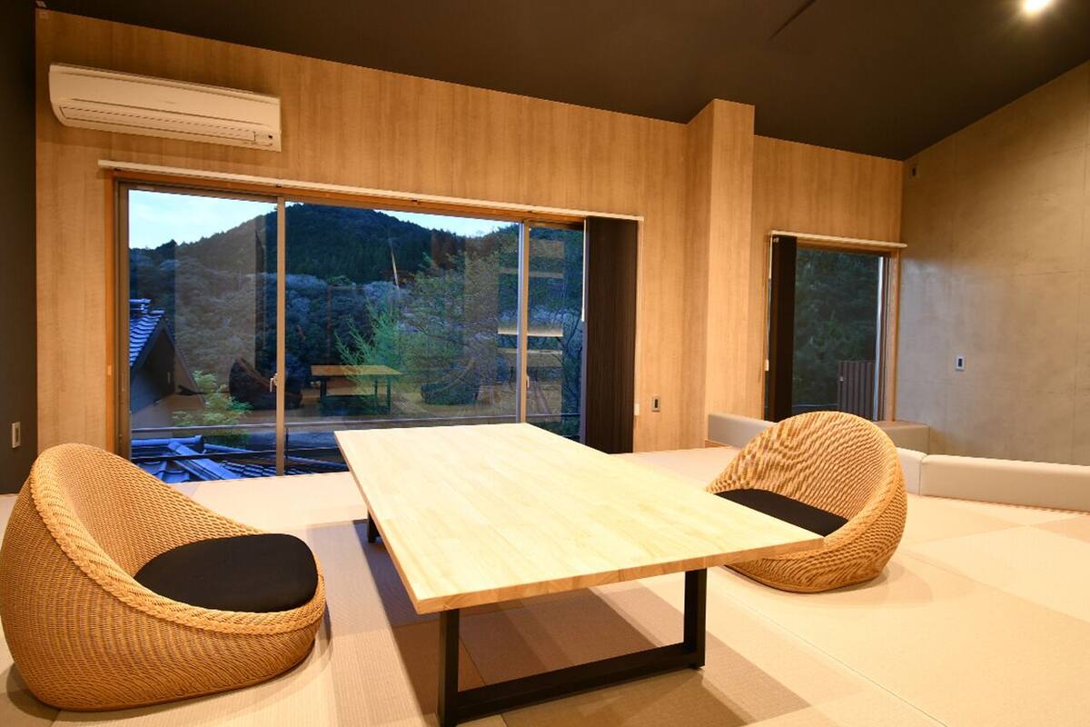 Une pièce moderne avec une grande table, deux chaises en osier et de grandes fenêtres donnant sur un paysage montagneux.