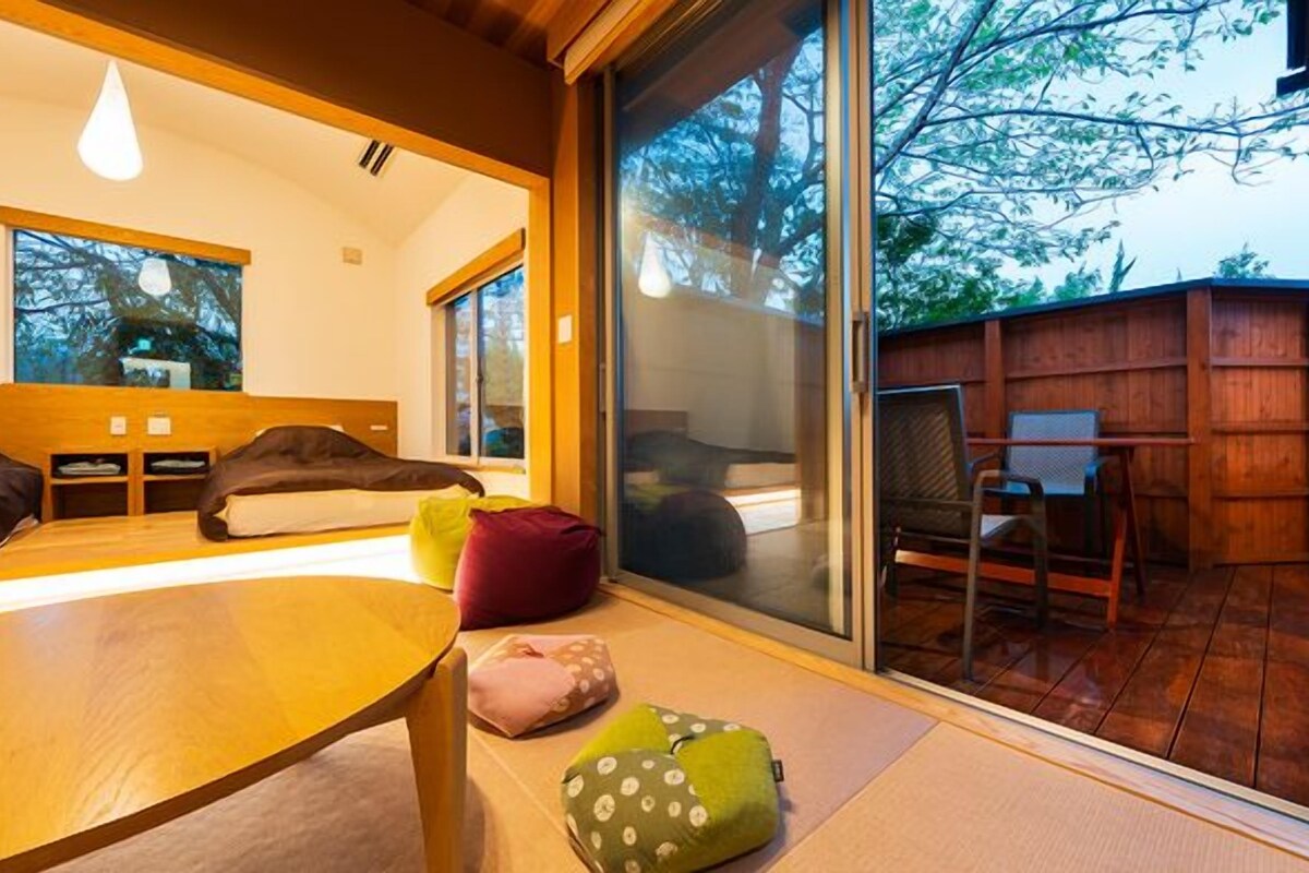 A modern room with a bed, a table, and a chair. There is a sliding glass door leading to an outdoor area with a wooden deck and a chair.