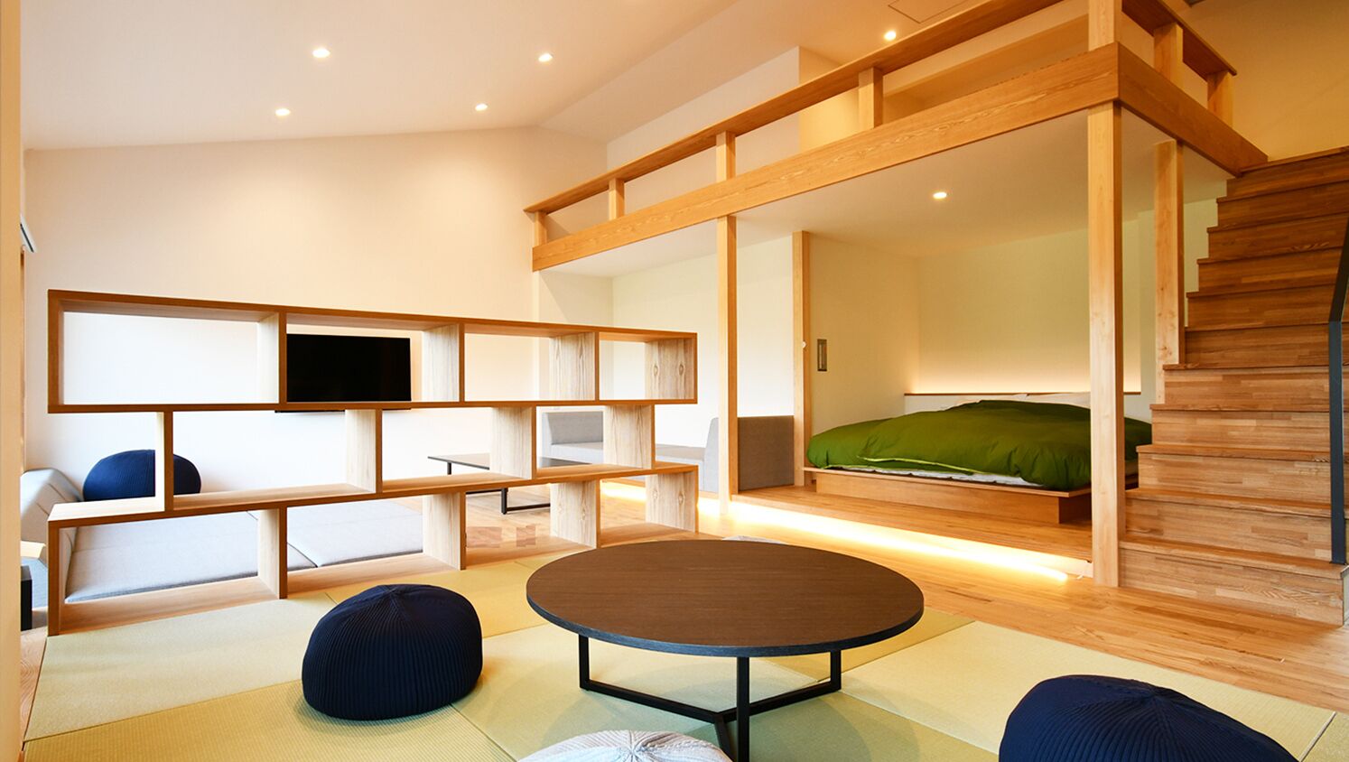 A modern room with a tatami mat floor, a round table, a sofa, a bed with a green mattress, and a TV on a wooden shelf.