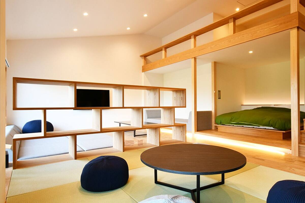 Une chambre moderne avec un sol recouvert de tatamis, une table ronde, un canapé, un lit avec un matelas vert et un téléviseur posé sur une étagère en bois.