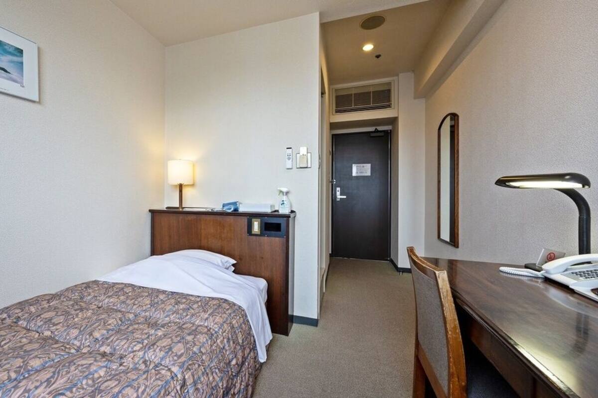 Habitación de hotel con cama, escritorio con lámpara, teléfono y puerta que da a un pasillo.