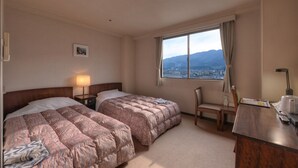 Camera d'albergo con due letti, una scrivania, una sedia, una lampada e una finestra vista città e montagne.