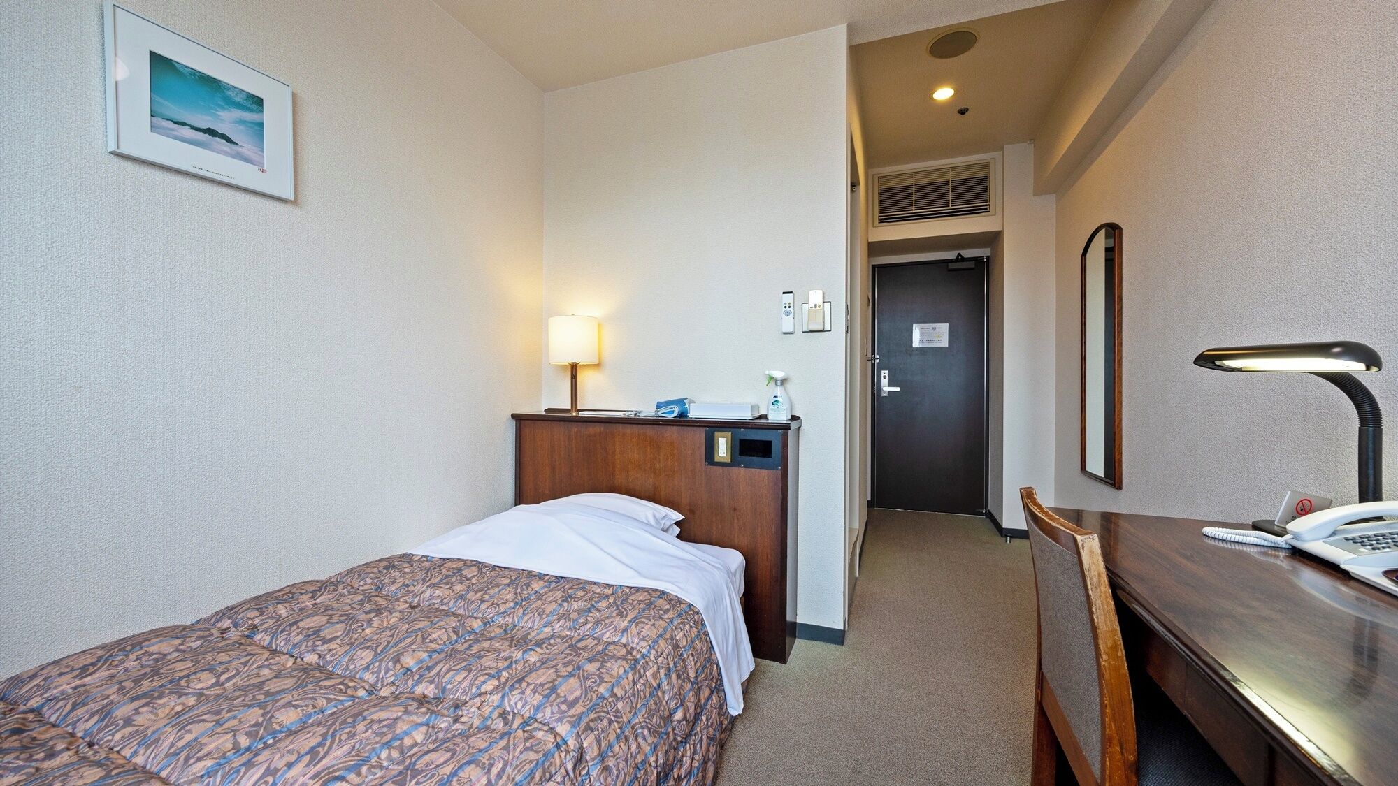 A hotel room with a bed, a desk, a lamp, a phone, and a door leading to a hallway.