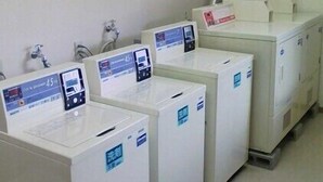 Een rij witte wasmachines met digitale displays, een in de muur gemonteerde wasbak en een ventilatiesysteem.