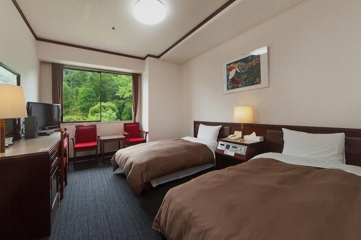 Een hotelkamer met twee bedden, een bureau met een computer, een stoel, een raam met uitzicht op groen, en een schilderij aan de muur.