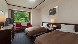 Een hotelkamer met twee bedden, een bureau met een computer, een stoel, een raam met uitzicht op groen, en een schilderij aan de muur.