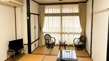 A small room with tatami mats, two chairs, a table, a TV, and a window with curtains.