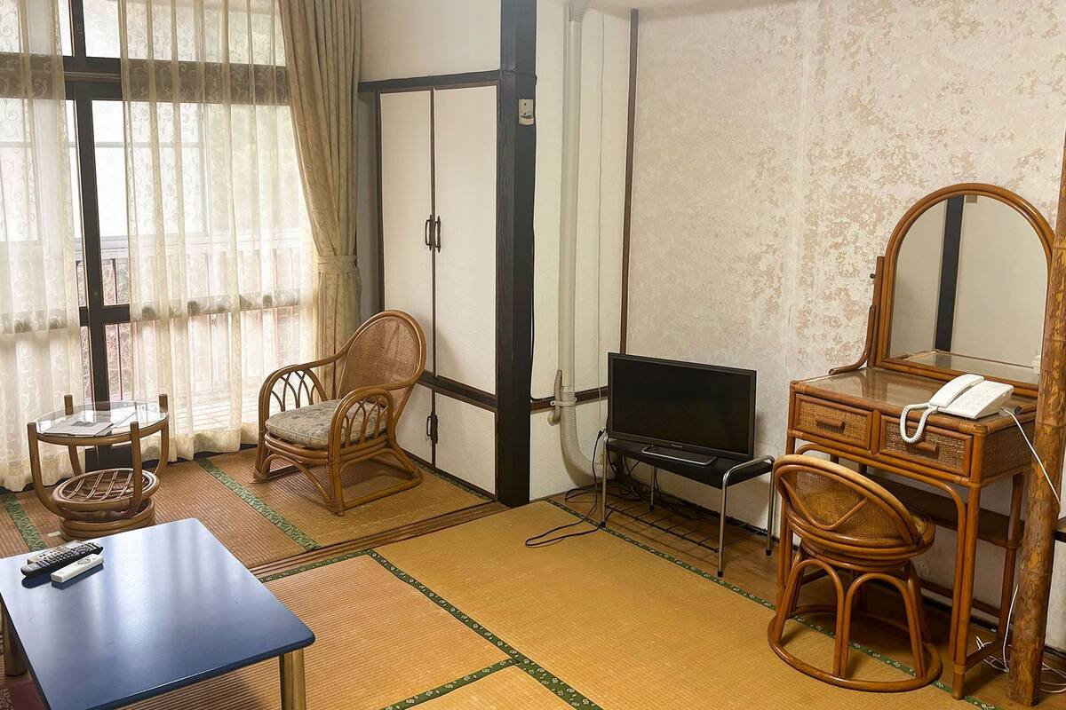 Ein Zimmer mit Tatami-Matten, einem Fernseher, einem Schreibtisch mit Telefon und einem traditionellen japanischen Sessel.