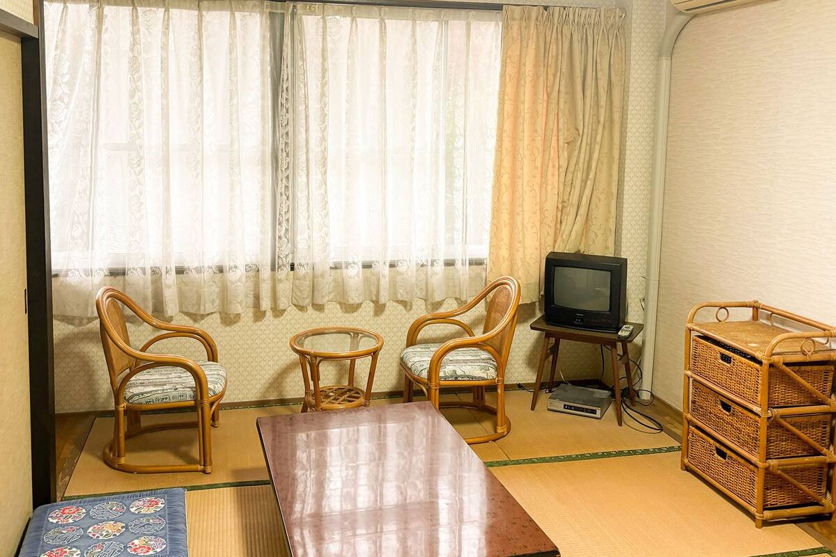 Ein Zimmer mit zwei Holzsesseln, einem kleinen Tisch, einem Fernseher auf einem Standfuß und einem Korbschrank.