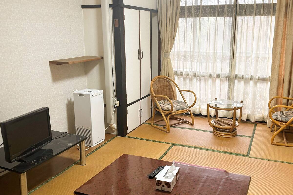 Ein Zimmer mit Fernseher, Klimaanlage, zwei Korbstühlen, einem kleinen Tisch und einem Boden mit Tatami-Matten.