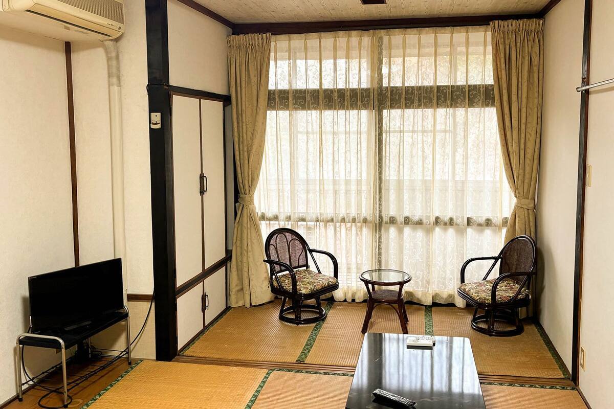 Ein Zimmer mit Tatami-Böden, einem Fernseher, zwei Stühlen, einem kleinen Tisch und einem Fenster mit durchscheinenden Vorhängen.