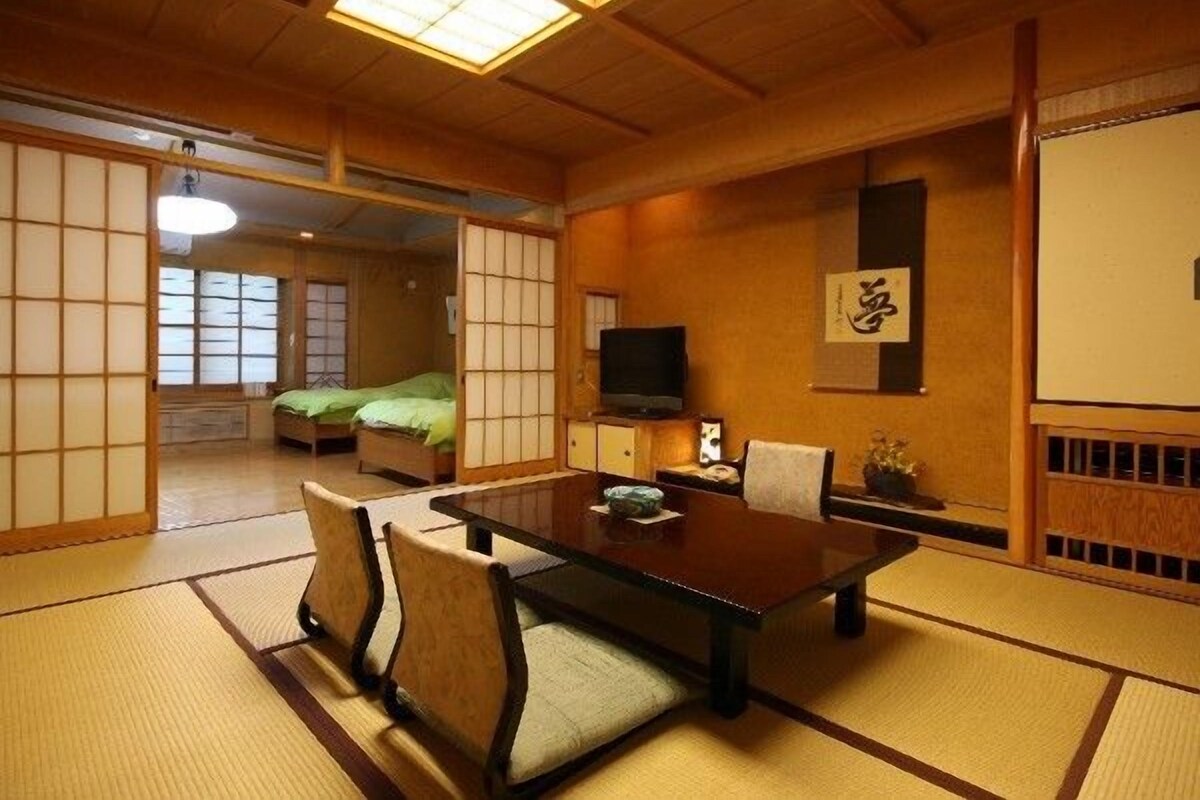 Una habitación japonesa tradicional con suelo de tatami, una mesa baja con sillas, un televisor y puertas correderas.