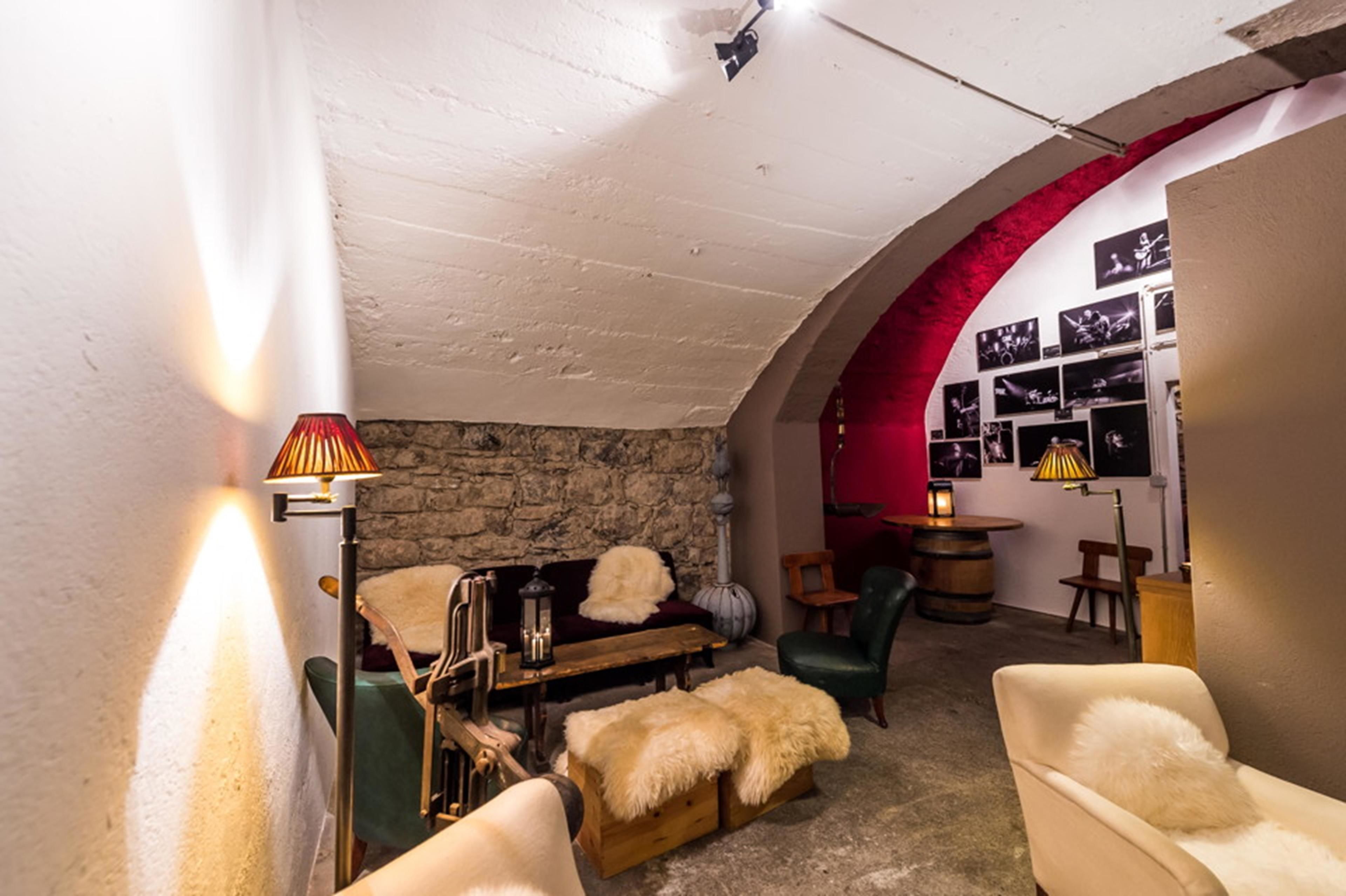 A cozy room with a stone wall, a wooden bench, and a green chair. There is a lamp on the left and a shelf with framed pictures on the right.
