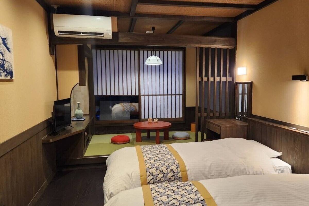 A hotel room with a bed, a television, a small table, a window with vertical blinds, and a wall-mounted air conditioner.