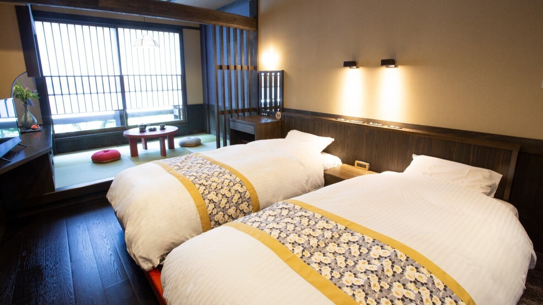 Habitación de hotel con dos camas, una mesita, una silla y una ventana con persianas.