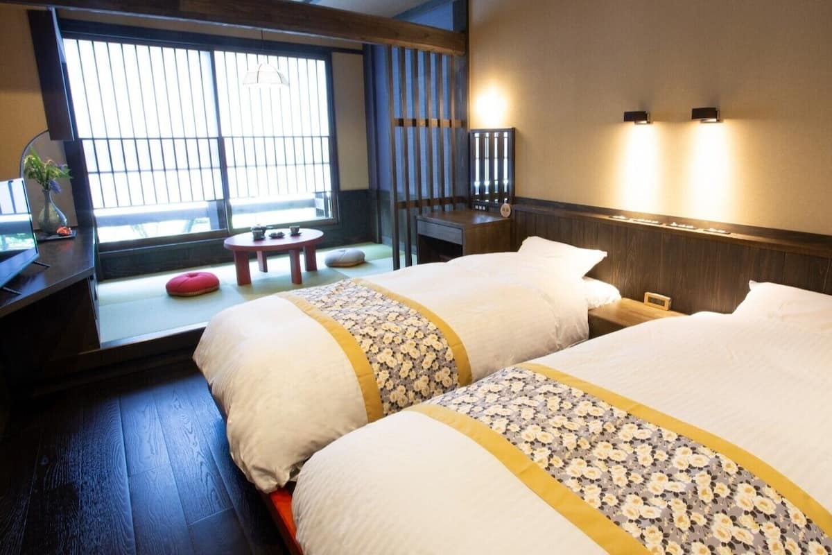 A hotel room with two beds, a small table, a chair, and a window with blinds.