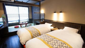A hotel room with two beds, a small table, a bench, and a window with blinds.