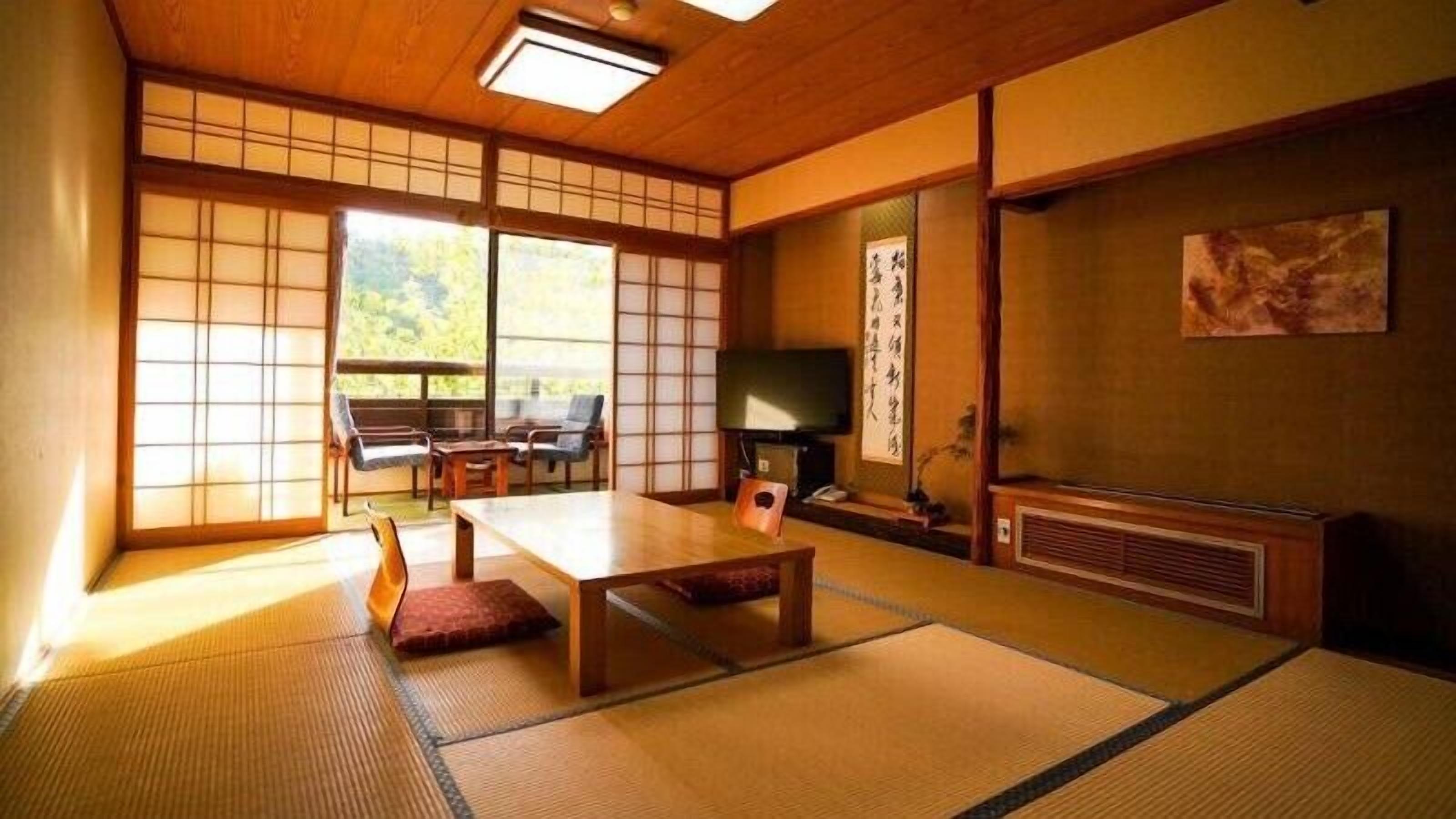 Una habitación japonesa tradicional con suelo de tatami, una mesa baja, sillas, un televisor y puertas correderas.