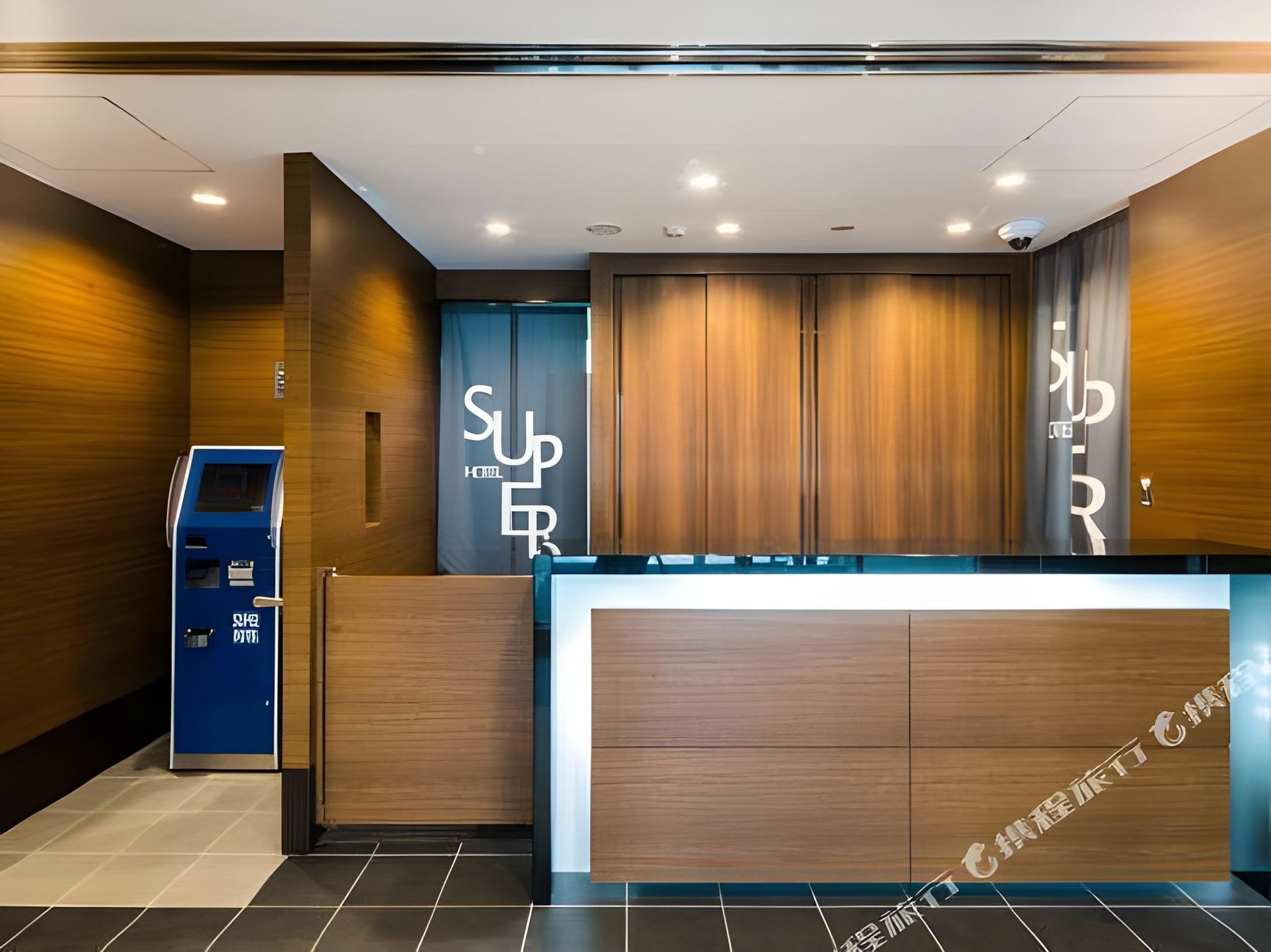 A hotel reception area with a wooden reception desk, a blue information kiosk, and a large sign with the hotel's name.